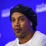 Ronaldinho, znany z imponującego wzrostu i uśmiechu, w czapce i białej koszulce na tle niebieskiej sceny.