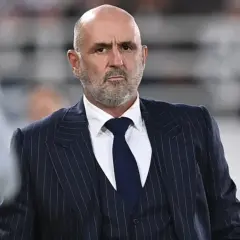 Łysy mężczyzna z brodą, ubrany w granatowy garnitur w paski i białą koszulę, to polski trener.