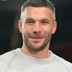 Uśmiechnięty Lukas Podolski w szarej bluzie. Jego kariera piłkarska i biznesowa zbudowały imponujący majątek.