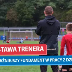 Trener piłki nożnej obserwuje młodych zawodników na boisku. Jego postawa jest kluczowa w pracy z dziećmi.
