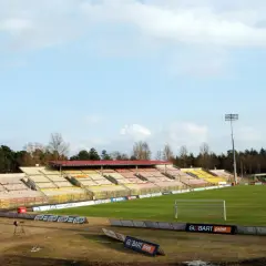 Puste trybuny starego stadionu Jagiellonii, z zieloną murawą i bramką. W tle las i niebo.
