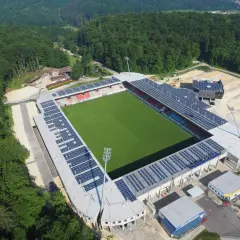 Widok z lotu ptaka na zieloną murawę i trybuny stadionu, otoczonego lasem. To najmniejszy stadion na świecie, z panelami słonecznymi na dachu.