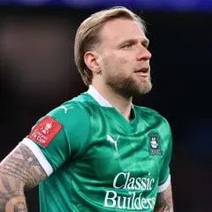 Piłkarz w zielonej koszulce z herbem klubu i naszywką FA Cup. Widać tatuaże na ramieniu. To właśnie tam gra puchacz.