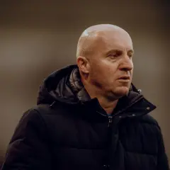 Nowy trener Widzewa, łysy mężczyzna w czarnej kurtce, patrzy w bok z zamyśloną miną.