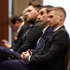 Mężczyźni w garniturach siedzą na sali, słuchając wykładu. Jeden z nich, z krótką fryzurą, wygląda na skupionego, być może uczestnicząc w szkoleniu UEFA Pro.