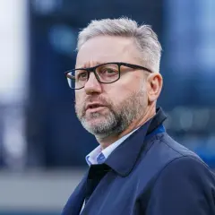 Mężczyzna w okularach, z siwą brodą i włosami, ubrany w ciemną kurtkę. Wygląda jak trener u21, skupiony na boisku.
