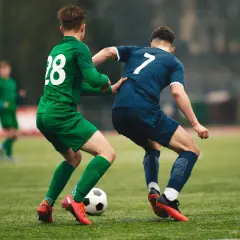 Piłkarze walczą o piłkę. Jeden w zielonej koszulce z numerem 28, drugi w granatowej z numerem 7. Nogi graczy są w ruchu, a boisko wygląda na lekko spalony od słońca.