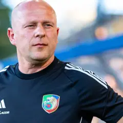 Łysy mężczyzna w czarnej koszulce z herbem Miedzi Legnica. Trener Miedzi Legnica w skupieniu obserwuje boisko.