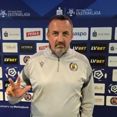 Trener Motoru Lublin z trzema palcami uniesionymi w geście zwycięstwa na tle sponsorów klubu.