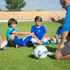 Trenerzy piłki nożnej instruują młodych zawodników siedzących na murawie.