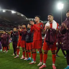 Piłkarze w czerwonych strojach, być może reprezentacja Chin, klaszczą na boisku. W tle widać trybuny i światła stadionu.