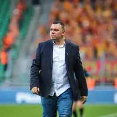Trener Jagiellonii w eleganckim garniturze i dżinsach, na tle trybun stadionu wypełnionych kibicami.