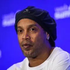 Ronaldinho, znany z imponującego wzrostu i uśmiechu, w czapce i białej koszulce na tle niebieskiej sceny.