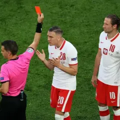 Sędzia pokazuje czerwoną kartkę, która wyklucza zawodnika z gry. Piłkarz z numerem 20 protestuje, a obok stoi jego kolega z numerem 10.