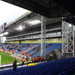 Trybuna stadionu Selhurst Park z niebieskimi i czerwonymi siedzeniami, zieloną murawą i tablicą wyników.