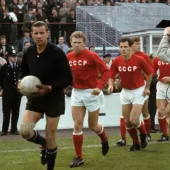 Bramkarz w czarnym stroju trzyma piłkę, obok niego biegną zawodnicy w czerwonych koszulkach z napisem CCCP. Kamera filmowa rejestruje mecz.