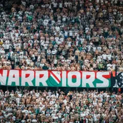 Tłum kibiców Legii Warszawa, z transparentem "WARRIORS", wspiera swoją drużynę.