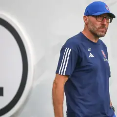 Marek Papszun w dresie Legii Warszawa, z charakterystycznym logo klubu na piersi, stoi obok wielkiej litery "L".