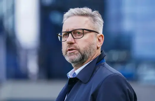 Mężczyzna w okularach, z siwą brodą i włosami, ubrany w ciemną kurtkę. Wygląda jak trener u21, skupiony na boisku.