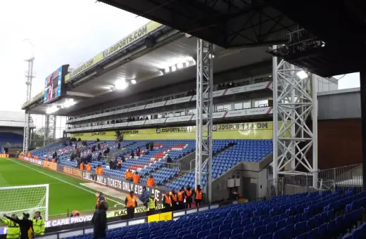 Trybuna stadionu Selhurst Park z niebieskimi i czerwonymi siedzeniami, zieloną murawą i tablicą wyników.