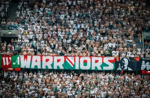 Tłum kibiców Legii Warszawa, z transparentem "WARRIORS", wspiera swoją drużynę.