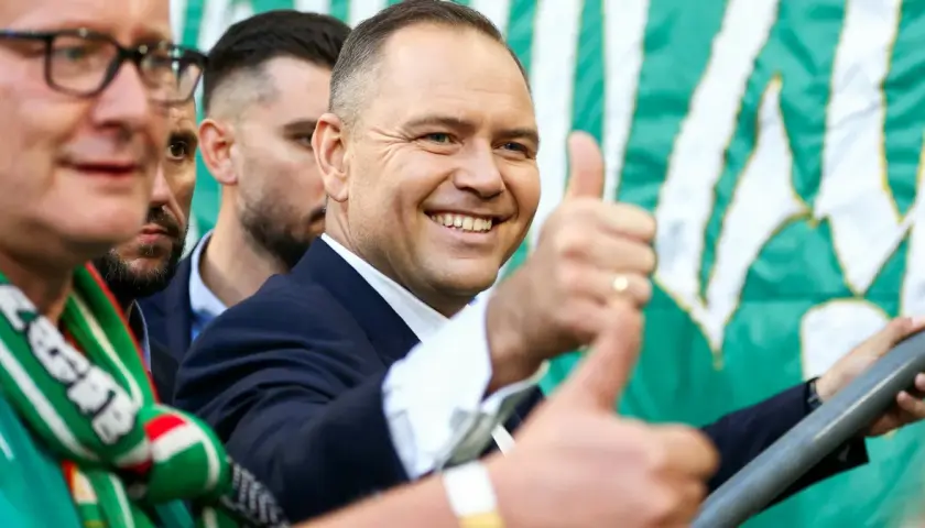 Uśmiechnięty mężczyzna w garniturze, z kciukiem w górze, na tle zielono-białej flagi. Widać kibiców Lechii Gdańsk.