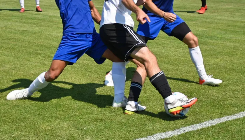 Piłkarze walczą o piłkę na boisku. Dwóch obrońców w niebieskich strojach próbuje odebrać futbolówkę zawodnikowi w białej koszulce.