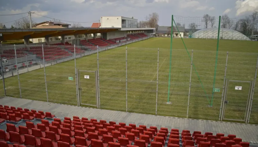 Puste trybuny i zielona murawa na stadionie w Niepołomicach. W tle budynek klubowy i balon nad boiskiem.