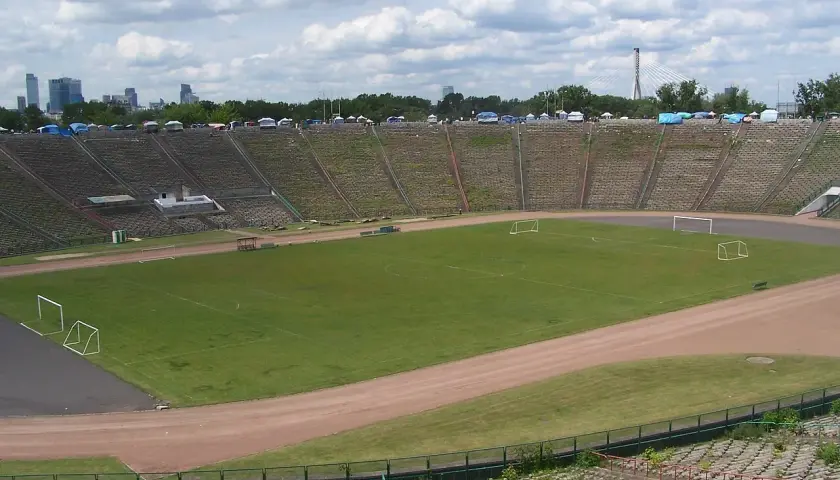Stadion z PRL z murawą, bramkami i trybunami, w tle panorama Warszawy.
