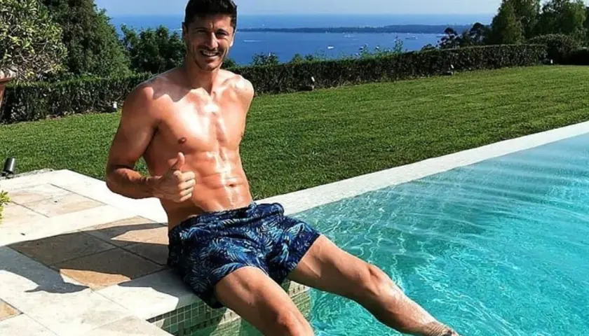 Robert Lewandowski w szortach z motywem liści siedzi na brzegu basenu, dając kciuk w górę. W tle błękitne morze i zieleń.