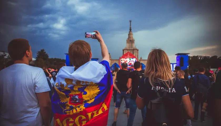 Młody kibic z flagą Rosji, wspierający klub piłkarski z Moskwy, filmuje telefonem wydarzenie przed monumentalnym budynkiem.