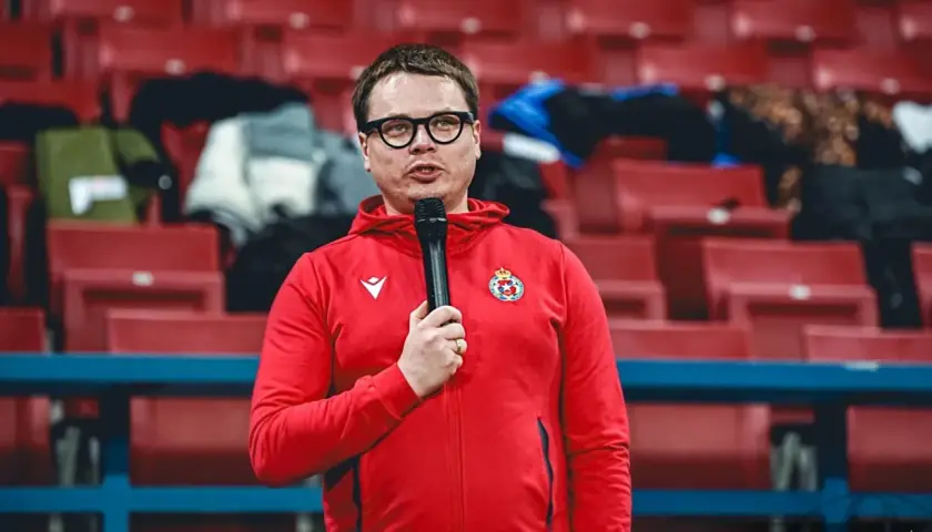 Właściciel Wisły Kraków w czerwonej bluzie z kapturem i mikrofonem, na tle pustych czerwonych trybun stadionu.
