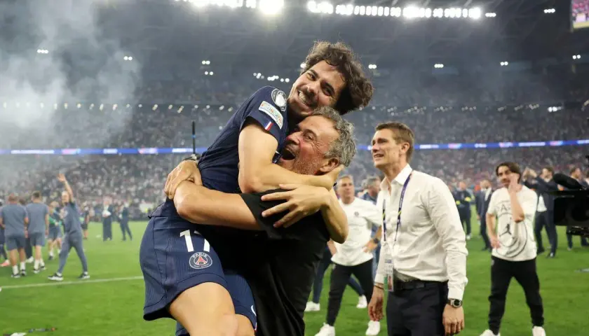 Radość piłkarza PSG obejmującego trenera. W tle tłum kibiców i stadion.