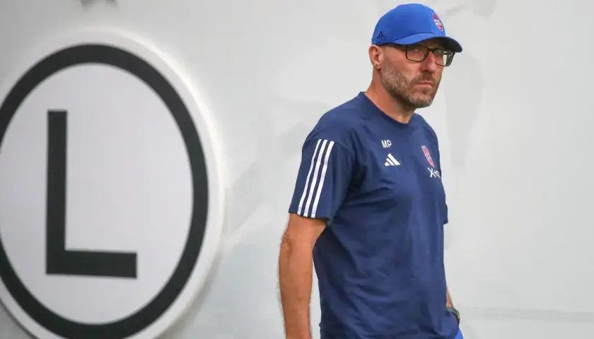 Marek Papszun w dresie Legii Warszawa, z charakterystycznym logo klubu na piersi, stoi obok wielkiej litery "L".