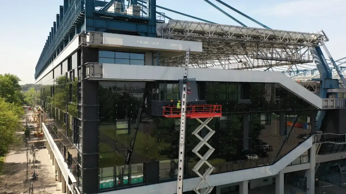 Prace przy stadionie Wisły po remoncie. Dwóch robotników na podnośniku montuje szklaną fasadę.