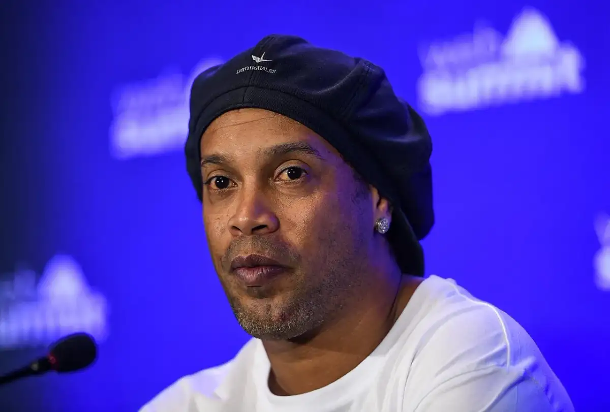 Ronaldinho, znany z imponującego wzrostu i uśmiechu, w czapce i białej koszulce na tle niebieskiej sceny.