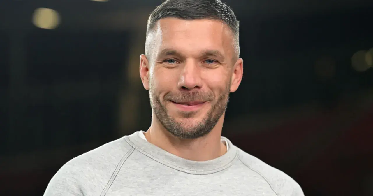 Uśmiechnięty Lukas Podolski w szarej bluzie. Jego kariera piłkarska i biznesowa zbudowały imponujący majątek.