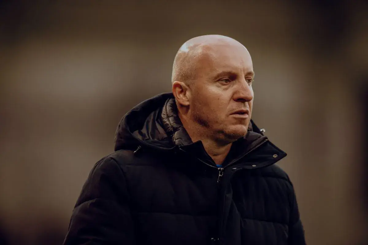 Nowy trener Widzewa, łysy mężczyzna w czarnej kurtce, patrzy w bok z zamyśloną miną.