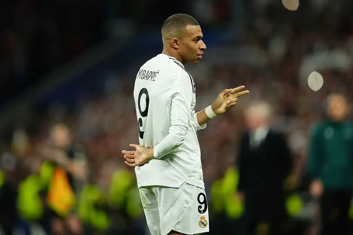Mbappe, w białej koszulce z numerem 9, gestykuluje na boisku. Jego wzrost robi wrażenie, a kibice śledzą każdy jego ruch.