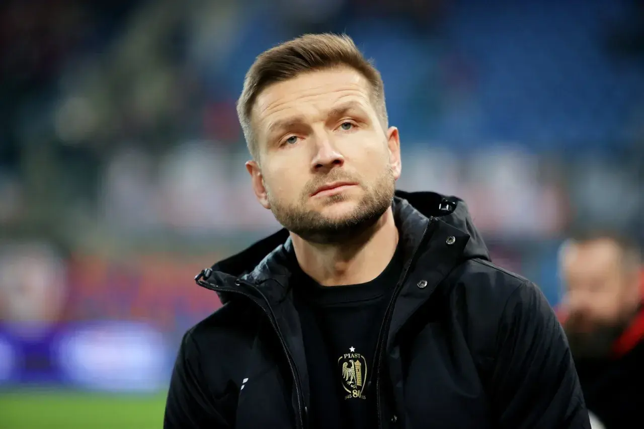 Mężczyzna w czarnej kurtce, z zarostem, w tle trybuny. To piast gliwice trener.