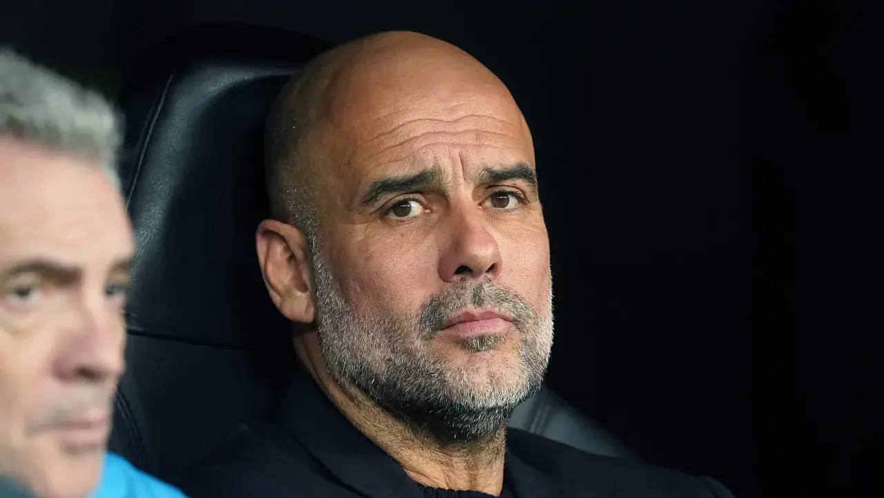 Pep Guardiola, zamyślony trener, siedzi na ławce, analizując grę.