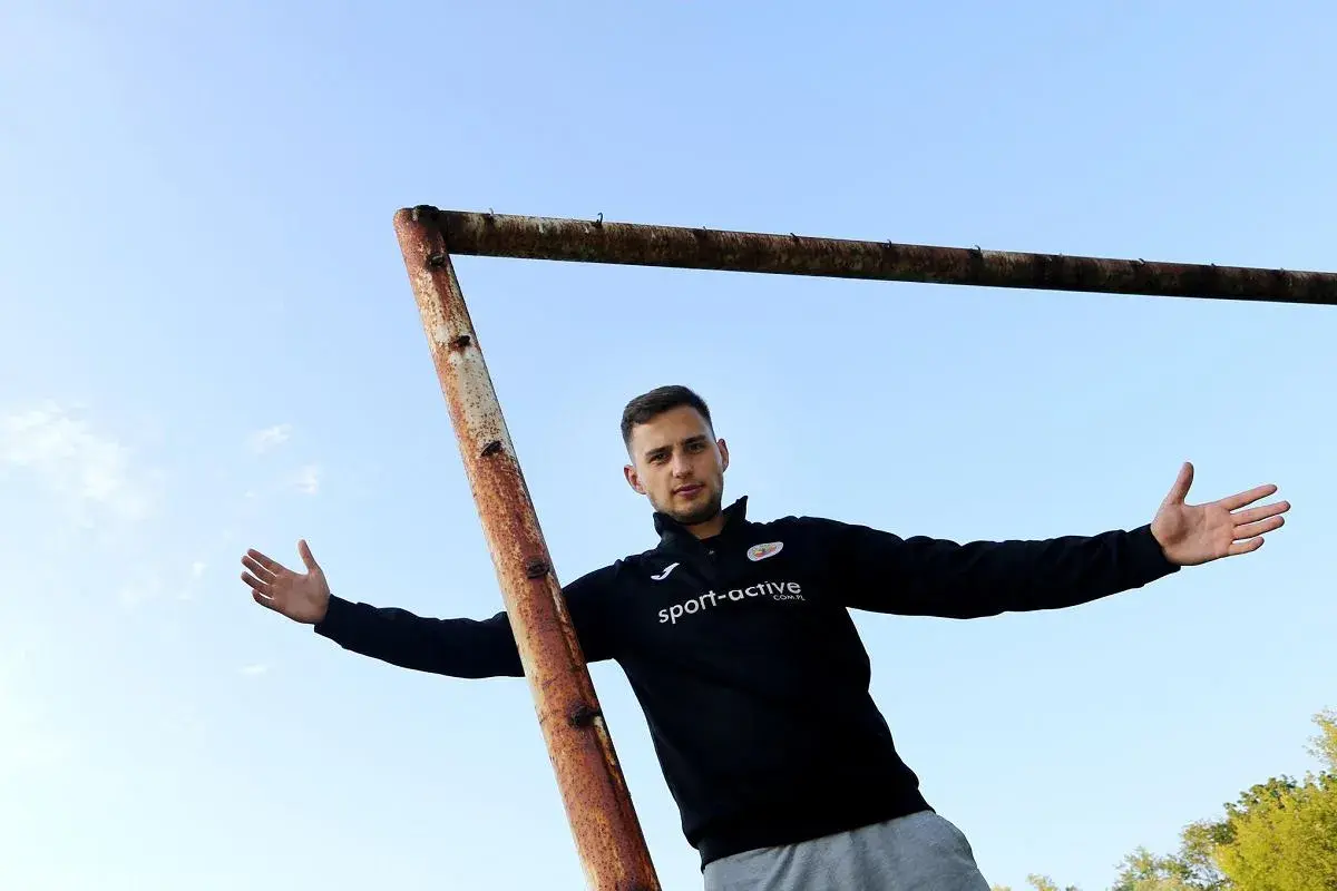 Młody piłkarz z Częstochowy, w czarnej bluzie z logo "sport-active", pozuje na tle nieba i zardzewiałej poprzeczki bramki.