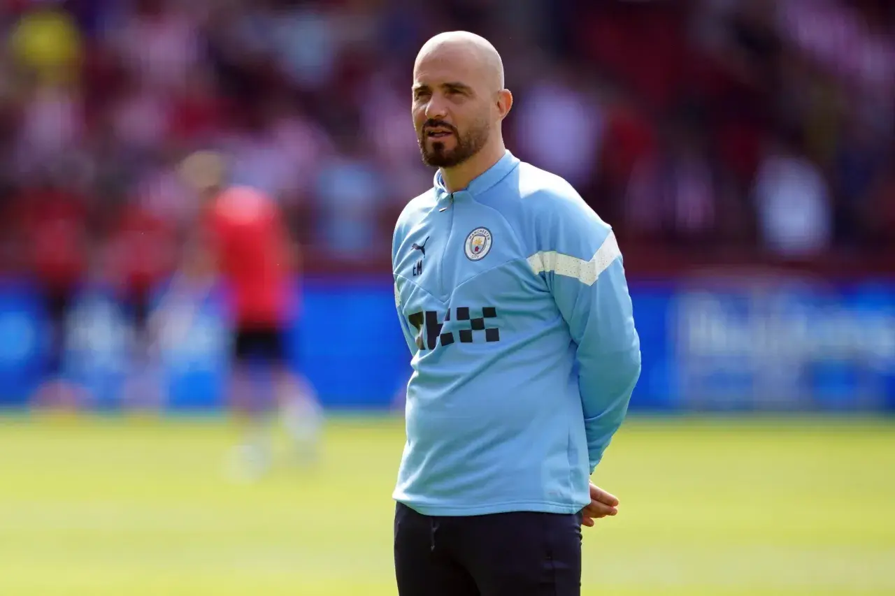 Łysy mężczyzna z brodą, trener Manchester City, w błękitnej bluzie treningowej, stoi na boisku.