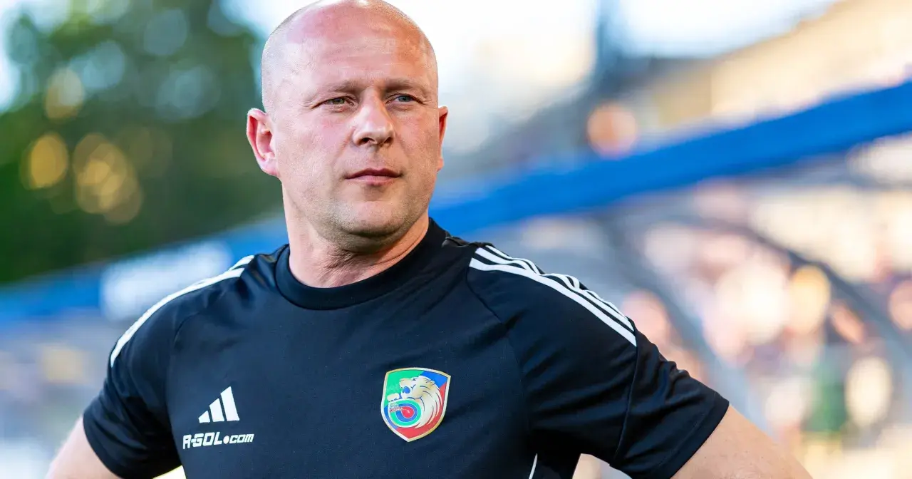 Łysy mężczyzna w czarnej koszulce z herbem Miedzi Legnica. Trener Miedzi Legnica w skupieniu obserwuje boisko.