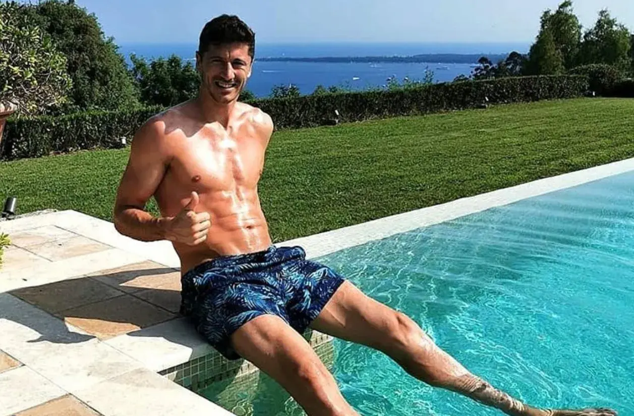 Robert Lewandowski w szortach z motywem liści siedzi na brzegu basenu, dając kciuk w górę. W tle błękitne morze i zieleń.