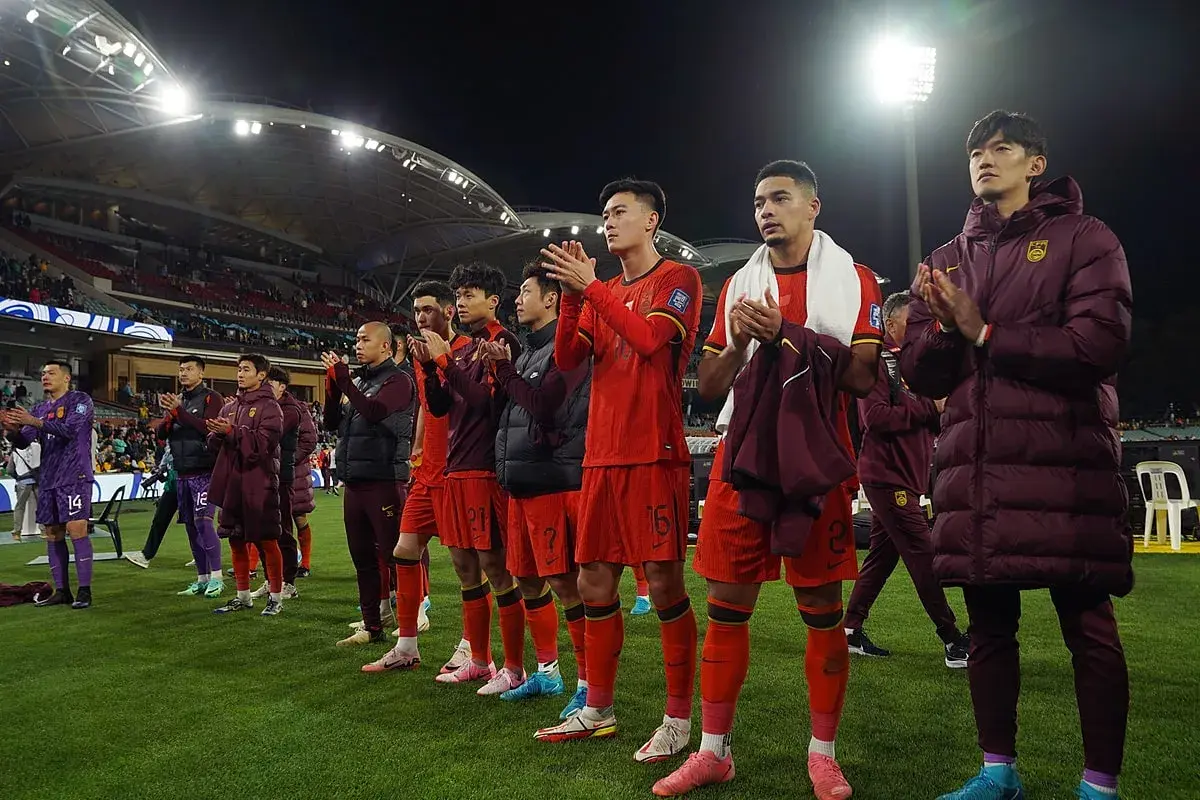 Piłkarze w czerwonych strojach, być może reprezentacja Chin, klaszczą na boisku. W tle widać trybuny i światła stadionu.