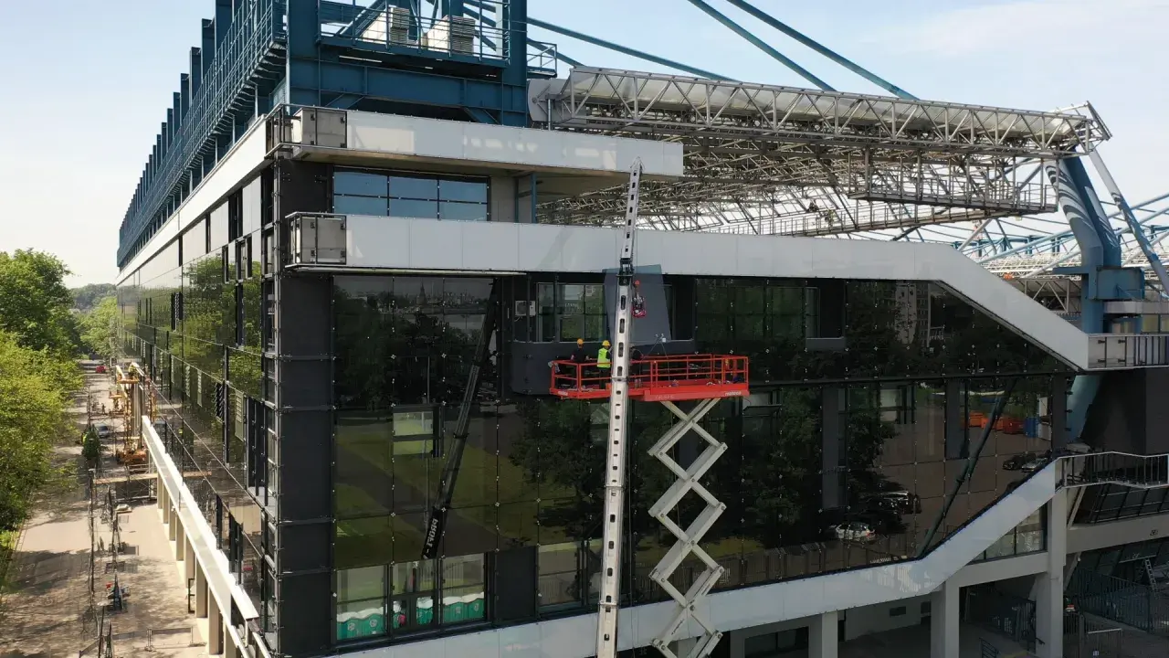 Prace przy stadionie Wisły po remoncie. Dwóch robotników na podnośniku montuje szklaną fasadę.
