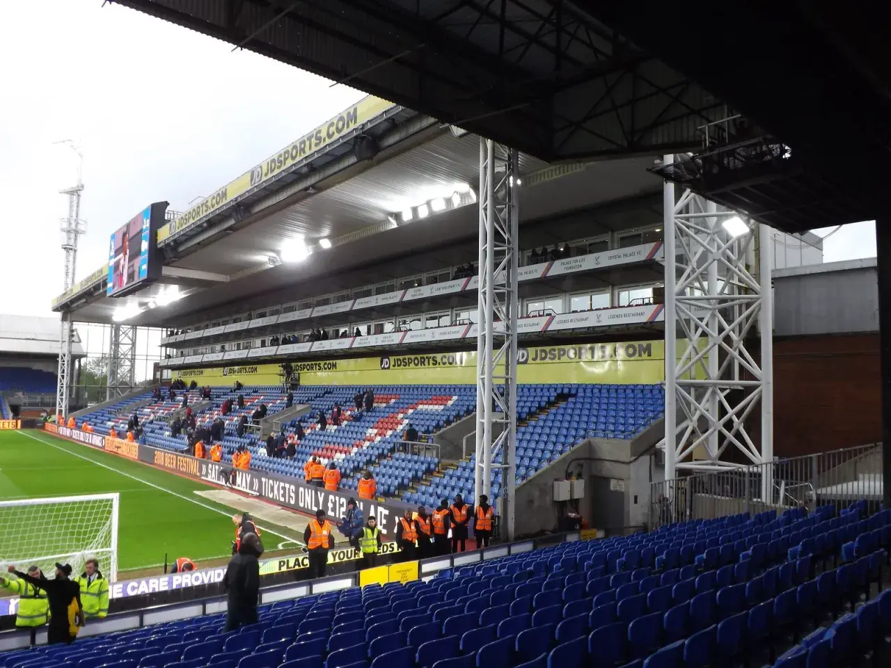Trybuna stadionu Selhurst Park z niebieskimi i czerwonymi siedzeniami, zieloną murawą i tablicą wyników.