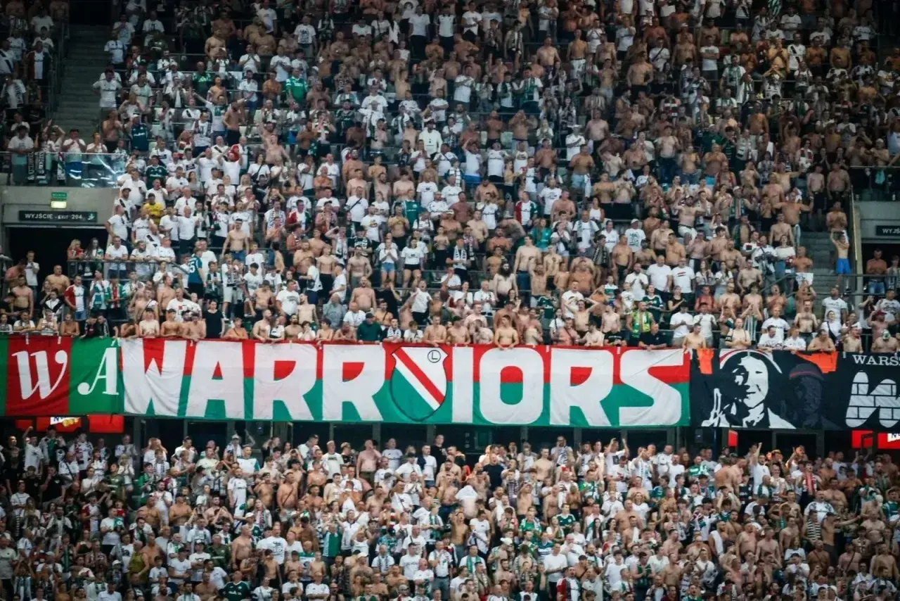 Tłum kibiców Legii Warszawa, z transparentem "WARRIORS", wspiera swoją drużynę.