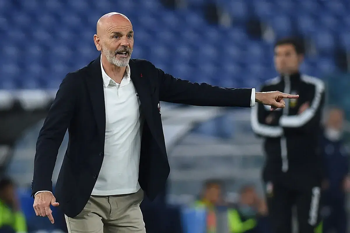 Trener Stefano Pioli w garniturze i białej koszulce polo wskazuje palcem, udzielając wskazówek na boisku.